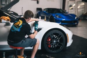 Image of a man applying ceramic coating to a car wheel at Empire Auto Spa. The man is wearing gloves and safety glasses, and he is using a spray bottle to apply the coating. The wheel is a shiny silver color.