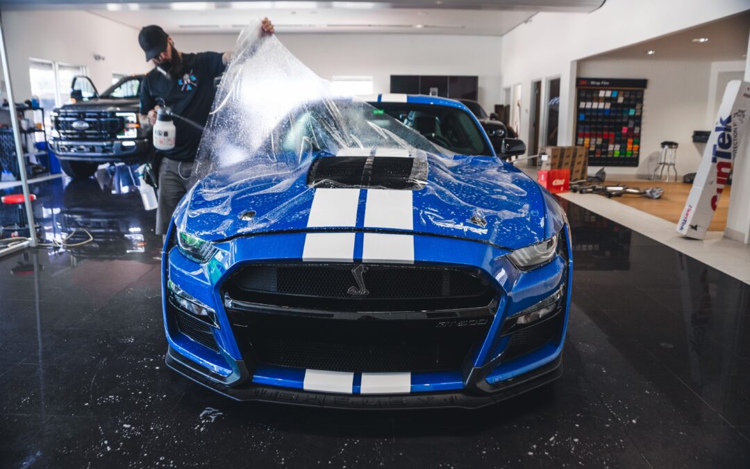 Image of a man wrapping a car in a garage. Paint Protection Film (PPF) being applied to a car. PPF is a clear, transparent film that protects the car's paint from scratches, chips, and other damage. It is a popular option for car owners who want to keep their car's paint looking new for years to come.